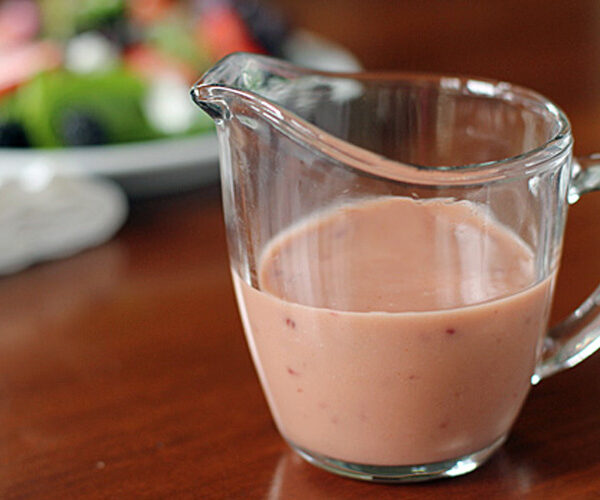 A glass pitcher filled with creamy pink salad dressing sits on a wooden table, with a blurred plate of salad in the background.