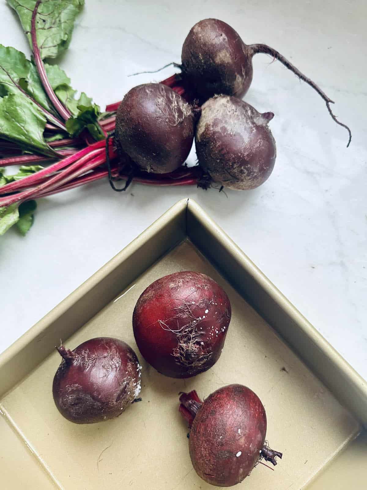 Four raw beets with stems and leaves rest on a light surface, while three smaller, cleaned beets sit inside a square, gold-colored tray nearby.