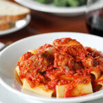 A white plate of rigatoni pasta topped with chunky tomato sauce and meat, with a glass of red wine and slices of bread in the background.