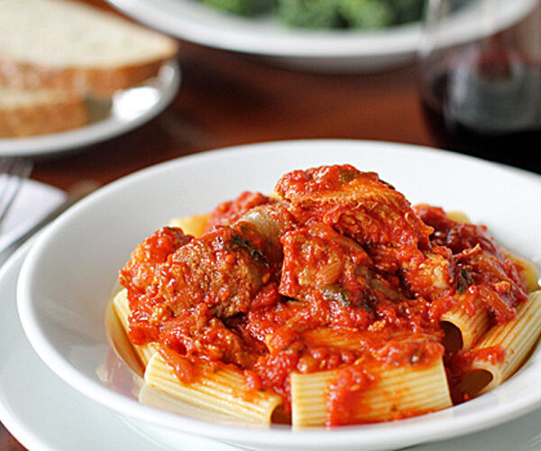 A white plate of rigatoni pasta topped with chunky tomato sauce and meat, with a glass of red wine and slices of bread in the background.