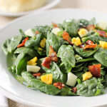 A white plate of fresh spinach salad topped with chopped hard-boiled eggs and crispy bacon pieces, placed on a light tablecloth with a fork and knife beside it.