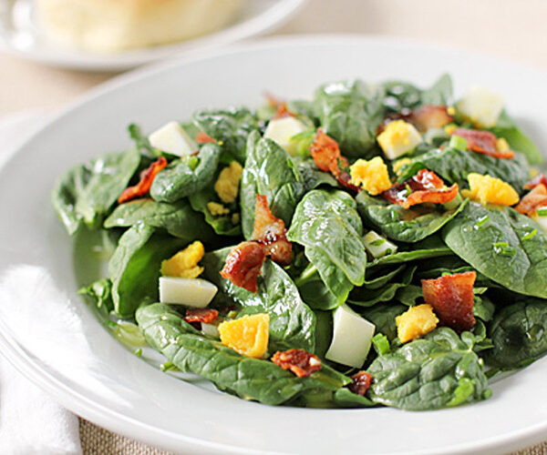 A white plate of fresh spinach salad topped with chopped hard-boiled eggs and crispy bacon pieces, placed on a light tablecloth with a fork and knife beside it.