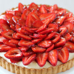 A strawberry tart with a golden crust, filled with cream and topped with neatly arranged slices of fresh, glossy strawberries.