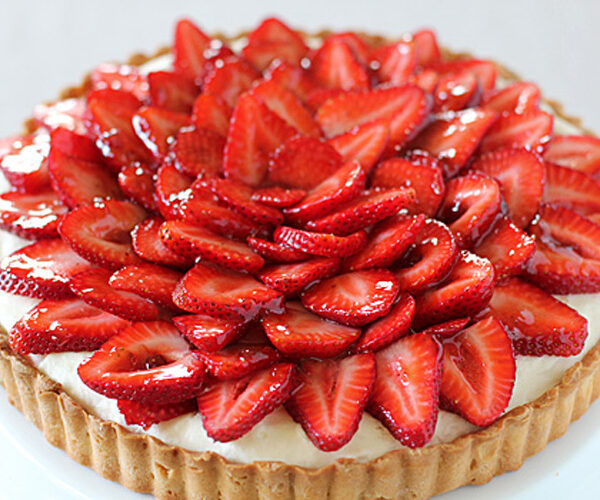 A strawberry tart with a golden crust, filled with cream and topped with neatly arranged slices of fresh, glossy strawberries.