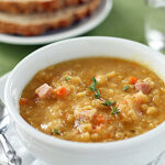 A bowl of thick split pea soup with diced ham and vegetables, garnished with fresh thyme. A spoon rests beside the bowl, with slices of whole-grain bread and a glass of water in the background.