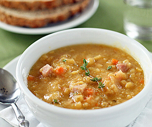 A bowl of thick split pea soup with diced ham and vegetables, garnished with fresh thyme. A spoon rests beside the bowl, with slices of whole-grain bread and a glass of water in the background.