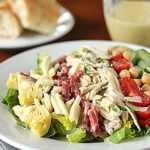 A plate of salad with sliced chicken, salami, artichoke hearts, cherry tomatoes, chickpeas, cheese, and lettuce. A fork and knife are beside the plate. Bread and a cup of dressing are in the background.