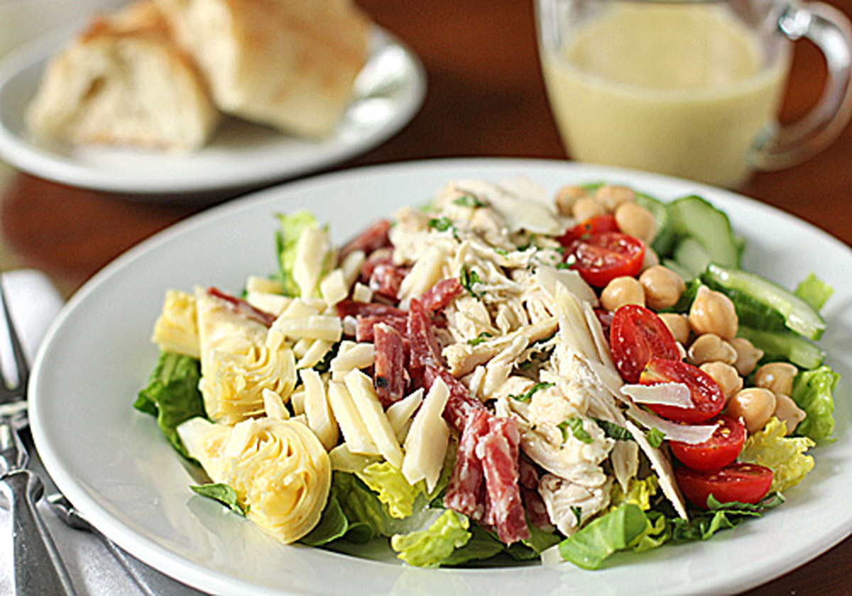 A plate of salad with sliced chicken, salami, artichoke hearts, cherry tomatoes, chickpeas, cheese, and lettuce. A fork and knife are beside the plate. Bread and a cup of dressing are in the background.
