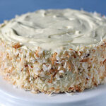 A round cake covered in creamy frosting and toasted shredded coconut on the sides sits on a white cake stand with a blurred blue background.