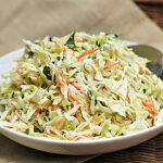 A white bowl filled with creamy coleslaw made of shredded cabbage, carrots, and herbs, with a spoon resting on the side. The bowl sits on a beige cloth with a wicker background.