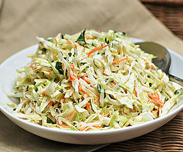 A white bowl filled with creamy coleslaw made of shredded cabbage, carrots, and herbs, with a spoon resting on the side. The bowl sits on a beige cloth with a wicker background.