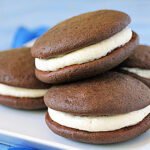 Four chocolate whoopie pies with white cream filling are stacked on a white plate, with a blue background.