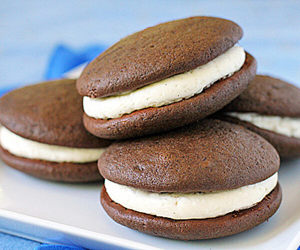 Four chocolate whoopie pies with white cream filling are stacked on a white plate, with a blue background.