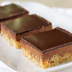 Three chocolate-topped dessert bars with a thick chocolate layer over a crumbly, light-colored base, arranged in a row on a white rectangular plate.