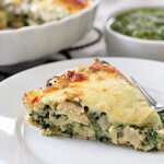 A slice of cheesy spinach and chicken quiche sits on a white plate with a fork. In the background, a baking dish with more quiche and a bowl of green sauce are visible.