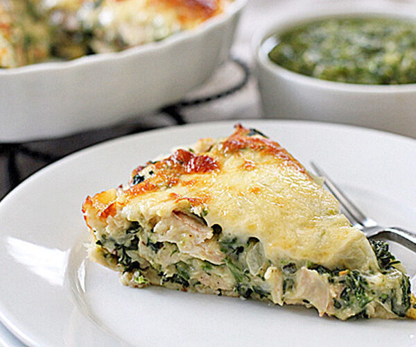 A slice of cheesy spinach and chicken quiche sits on a white plate with a fork. In the background, a baking dish with more quiche and a bowl of green sauce are visible.