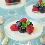 Glass dessert dishes filled with creamy panna cotta, each topped with fresh raspberries, blackberries, blueberries, and a small mint sprig, set on a light blue surface with silver spoons nearby.