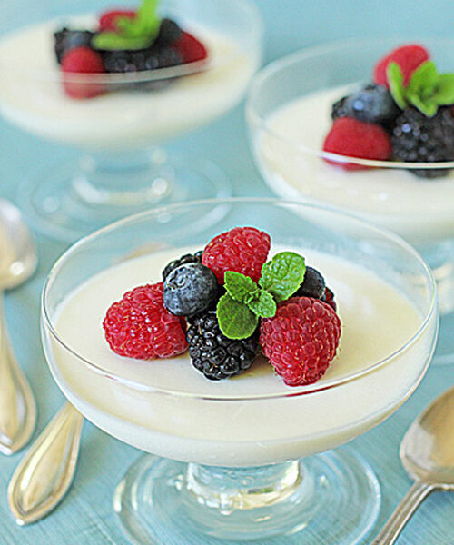 Glass dessert dishes filled with creamy panna cotta, each topped with fresh raspberries, blackberries, blueberries, and a small mint sprig, set on a light blue surface with silver spoons nearby.