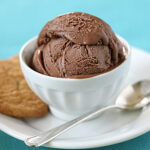A white bowl with two scoops of chocolate ice cream sits on a white plate, next to a cookie and a silver spoon, all placed on a blue surface.