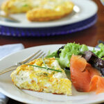 A plate with a slice of frittata, mixed greens, and smoked salmon, with a fork beside it. Another plate and a blue glass are in the blurred background.