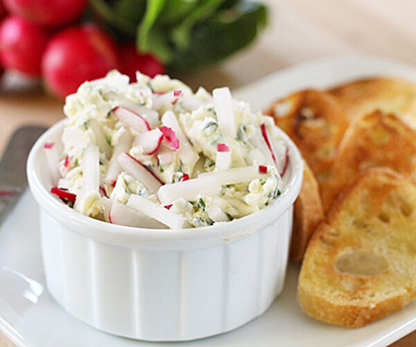 A white ramekin filled with a creamy dip garnished with chopped radishes and herbs, served on a plate with toasted baguette slices. Fresh radishes and greens are blurred in the background.