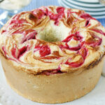 A round marble cake with swirls of raspberry on top sits on a white plate. A serving utensil is nearby, with plates and fresh raspberries in the background on a blue checkered tablecloth.