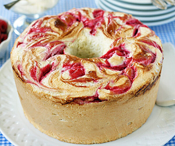 A round marble cake with swirls of raspberry on top sits on a white plate. A serving utensil is nearby, with plates and fresh raspberries in the background on a blue checkered tablecloth.
