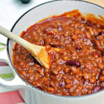 A white pot filled with thick, hearty chili made with beans and tomato sauce. A wooden spoon rests inside the pot, stirring the mixture. The pot sits on a striped cloth.