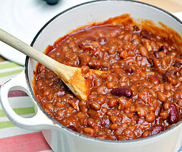 A white pot filled with thick, hearty chili made with beans and tomato sauce. A wooden spoon rests inside the pot, stirring the mixture. The pot sits on a striped cloth.