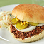 A sloppy joe sandwich on a white plate, topped with sliced onions, pickles, and mustard, with a side of coleslaw.