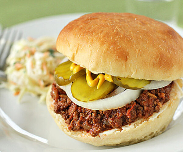 A sloppy joe sandwich on a white plate, topped with sliced onions, pickles, and mustard, with a side of coleslaw.