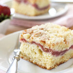 A square slice of crumbly coffee cake with a strawberry filling layer sits on a white plate with a fork. Fresh strawberries and another plate of cake are blurred in the background.