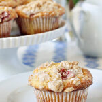 A crumb-topped muffin sits on a white plate, with more muffins displayed on a cake stand in the background. A teapot and blurred flowers are also visible.