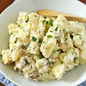 A bowl of creamy potato salad garnished with chopped parsley, served on a white plate with a wooden spoon, set on a blue cloth on a wooden table.