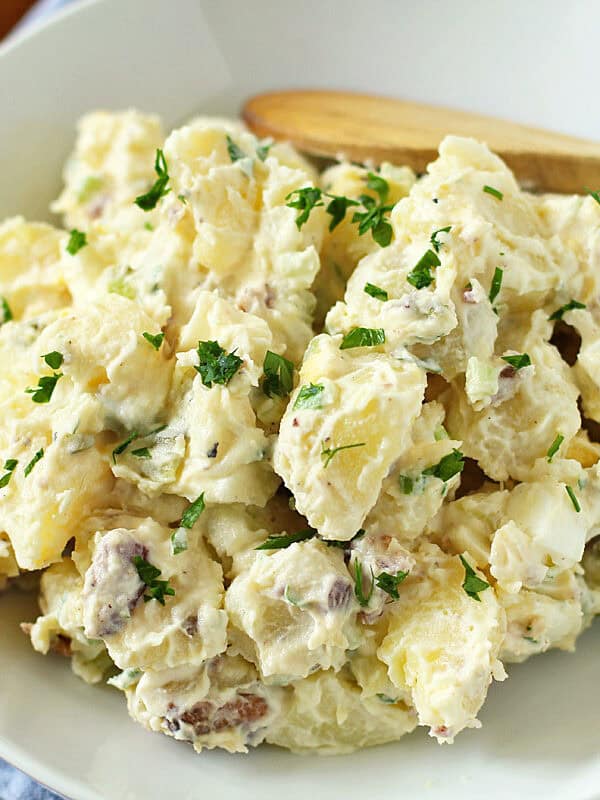 A bowl of creamy potato salad garnished with chopped parsley, served on a white plate with a wooden spoon, set on a blue cloth on a wooden table.
