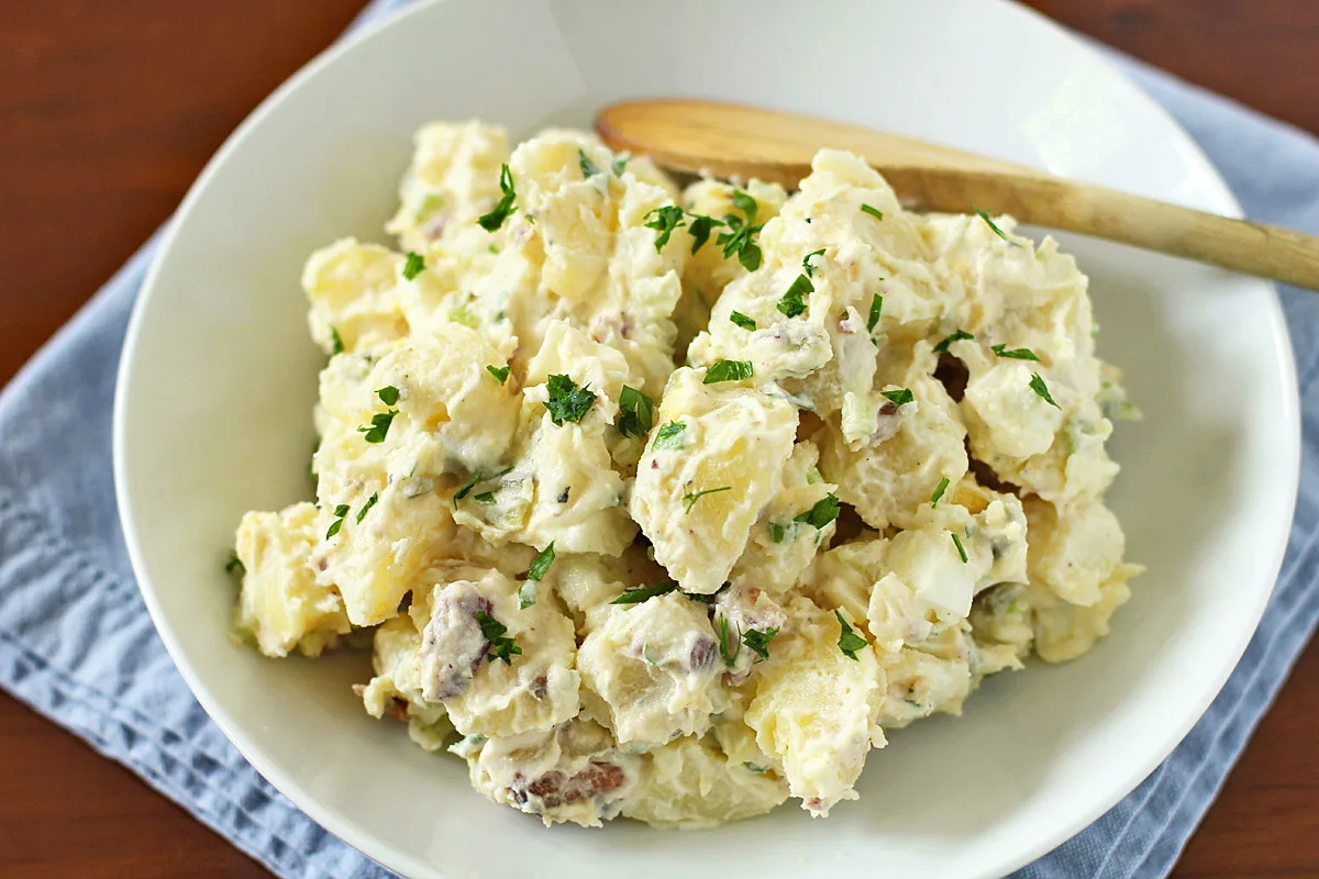 A bowl of creamy potato salad garnished with chopped parsley, served on a white plate with a wooden spoon, set on a blue cloth on a wooden table.