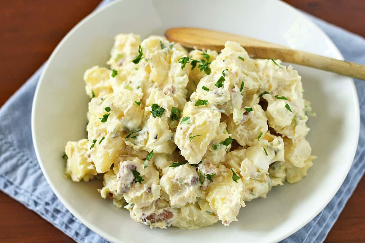 A bowl of creamy potato salad garnished with chopped parsley, served on a white plate with a wooden spoon, set on a blue cloth on a wooden table.