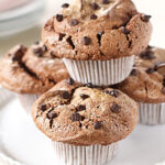 Three chocolate chip muffins in white paper liners are stacked on a white scalloped cake stand, with more muffins blurred in the background. The tops are cracked and sprinkled with mini chocolate chips.