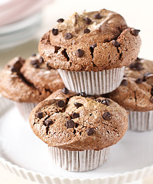 Three chocolate chip muffins in white paper liners are stacked on a white scalloped cake stand, with more muffins blurred in the background. The tops are cracked and sprinkled with mini chocolate chips.