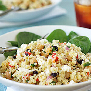 A white bowl filled with couscous salad featuring chickpeas, feta cheese, chopped vegetables, and herbs, served on a bed of fresh spinach leaves with a fork resting on the bowl. A glass of iced tea is in the background.