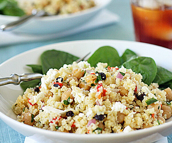 A white bowl filled with couscous salad featuring chickpeas, feta cheese, chopped vegetables, and herbs, served on a bed of fresh spinach leaves with a fork resting on the bowl. A glass of iced tea is in the background.