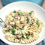 A person holds a white bowl filled with rotini pasta salad, featuring green peas, chunks of tuna, and diced vegetables, with a fork resting inside the bowl.