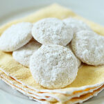 A group of round, powdered sugar-coated cookies arranged on a stack of yellow cloth napkins on a white plate.