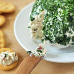 A wooden spreader lifts herbed cheese spread from a large cheese ball coated in chopped parsley, with round crackers arranged nearby on a wooden surface.
