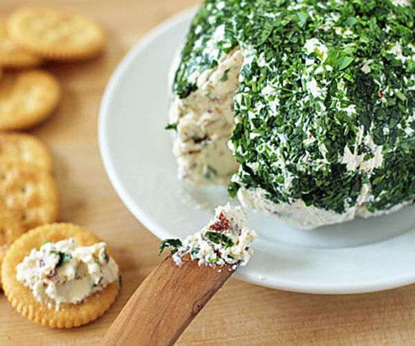 A wooden spreader lifts herbed cheese spread from a large cheese ball coated in chopped parsley, with round crackers arranged nearby on a wooden surface.