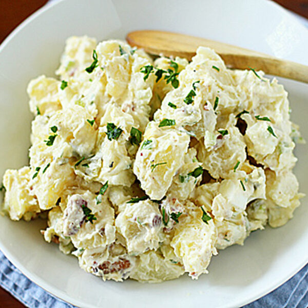 A white bowl filled with creamy potato salad topped with chopped parsley, with a wooden spoon resting on the side, placed on a blue cloth.
