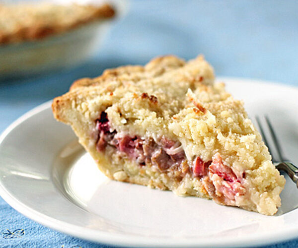 A slice of crumb-topped fruit pie with a golden crust is served on a white plate with a fork, with the rest of the pie visible in the background on a blue tablecloth.