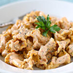 A plate of mafaldine pasta coated in a creamy sauce, garnished with fresh parsley and sprinkled with black pepper, with a fork resting on the side.
