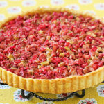 A baked rhubarb tart with a golden crust, topped with chopped pink and green rhubarb pieces, sits on a metal stand over a yellow floral tablecloth.
