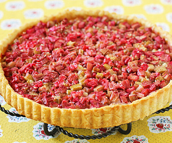 A baked rhubarb tart with a golden crust, topped with chopped pink and green rhubarb pieces, sits on a metal stand over a yellow floral tablecloth.